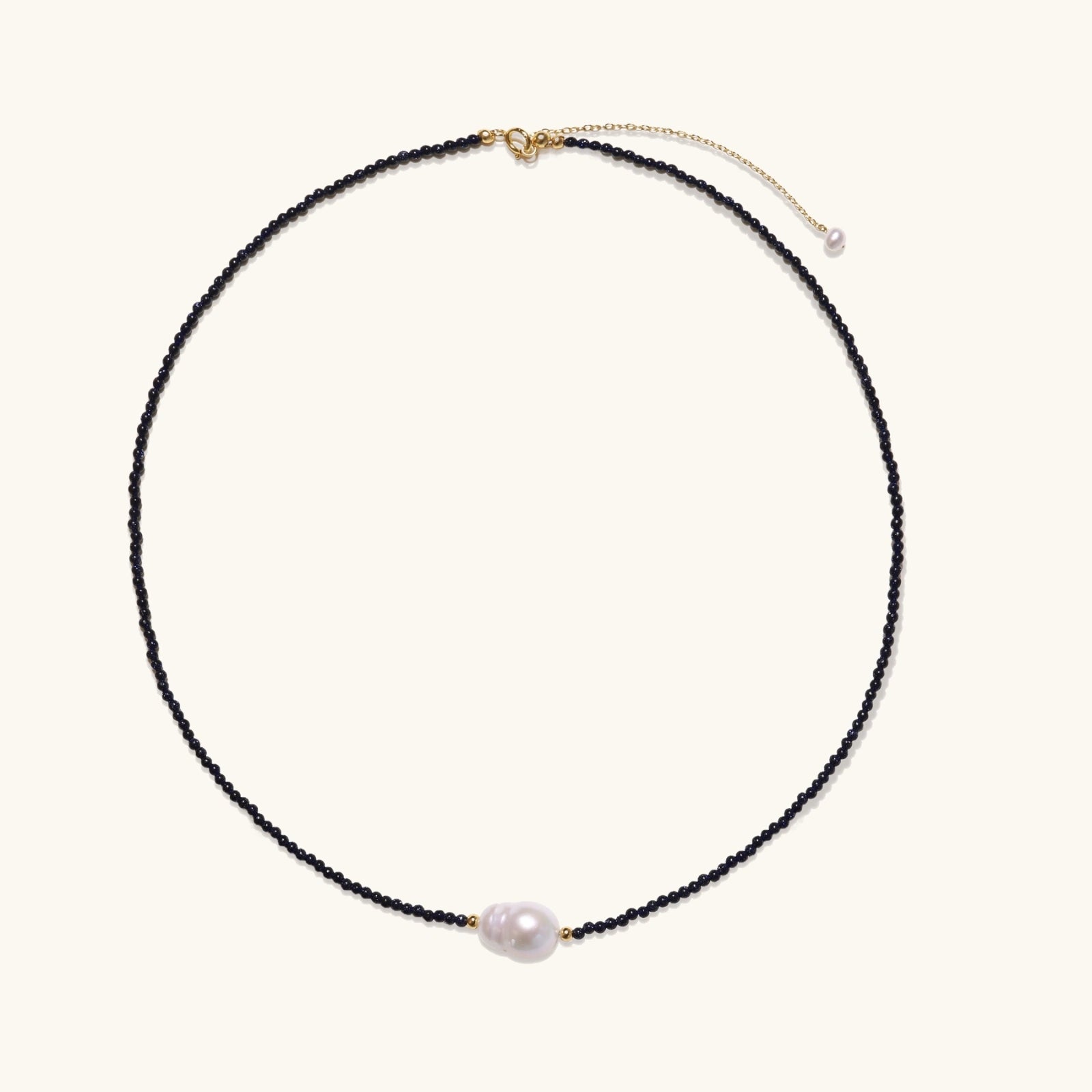 Delicate necklace with black blue sandstone beads and baroque pearl center on white background with gold accents.