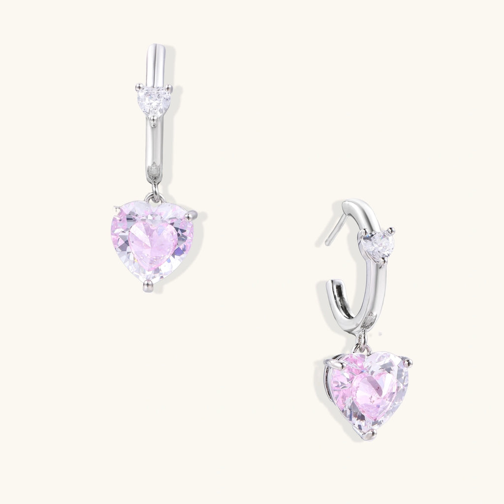 Silver huggie hoops with pink heart-cut zirconia pendants on white background, eco-friendly white gold plated brass