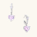 Silver huggie hoops with pink heart-cut zirconia pendants on white background, eco-friendly white gold plated brass