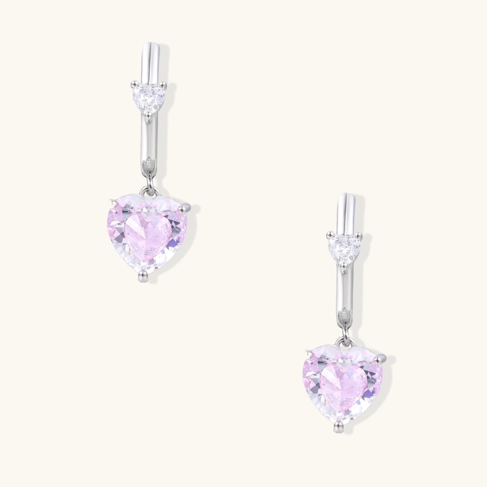Silver huggie hoops with pink heart-cut zirconia pendants on white background, eco-friendly white gold plated brass