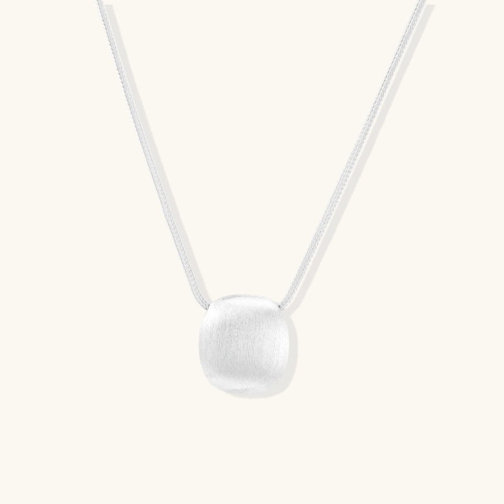 Minimalist brushed sterling silver dome pendant on delicate chain against cream background, showing matte textured finish